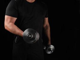 man in black clothes holds steel dumbbells in his hands, his muscles are tense photo