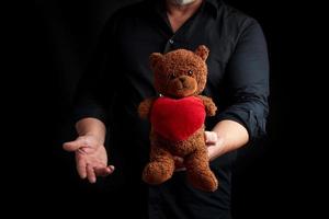 adult man in a black shirt holds a brown teddy bear with a red heart on a dark background photo