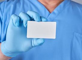 médico masculino con guantes de látex azules sostiene una tarjeta de visita de papel blanco en blanco foto