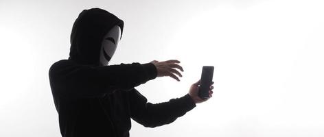 hacker anónimo y mascarilla con smartphone en la mano. hombre con camisa de capucha negra sosteniendo y usando un teléfono móvil con fondo blanco. representan el concepto de piratería informática o robo de datos personales foto