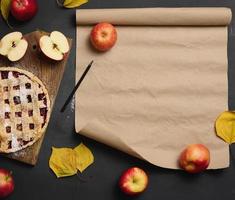 baked round traditional apple pie on a brown wooden board and fresh red apples. Nearby is a roll of brown paper for writing a recipe or menu, top view photo