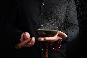 adult man in a black shirt rotates a wooden stick around a copper Tibetan bowl with water photo