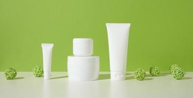 jar, empty white plastic tubes for cosmetics on a white table, green background. Packaging for cream, gel, serum, advertising and product promotion photo