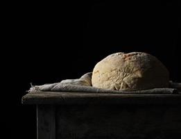 pan de trigo blanco redondo horneado sobre una toalla textil, mesa antigua de madera, fondo negro foto