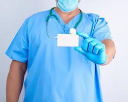 médico con guantes de látex estériles y uniforme azul tiene una tarjeta de visita blanca en blanco, foto