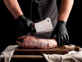 chef in black latex gloves holds a big knife and cuts into pieces raw rabbit meat on a brown wooden cutting board photo