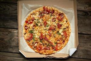 baked round pizza with smoked sausages, mushrooms, tomatoes, cheese and dill in an open cardboard box on a wooden table photo