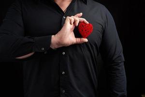 hombre adulto parado sobre un fondo oscuro con una camisa negra y sosteniendo un corazón rojo tallado cerca de su pecho foto