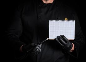 chef in black uniform and gloves is holding a blank notebook and a black wooden pencil photo