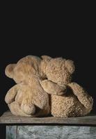 two old brown teddy bears sit hugging on a wooden surface photo