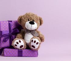 lindo oso de peluche marrón y caja envuelta en papel y cinta de seda sobre un fondo morado. premio y felicitaciones foto