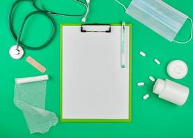 soporte de papel con sábanas blancas vacías, estetoscopio médico, pastillas sobre un fondo verde foto