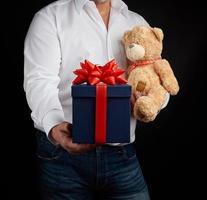 adult man in a white shirt holds a blue square box tied with a red ribbon and brown teddy bear photo