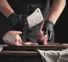 chef in black latex gloves holds a big knife and cuts into pieces raw rabbit meat photo