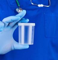 doctor in blue uniform and latex gloves is holding an empty plastic container for taking urine samples photo