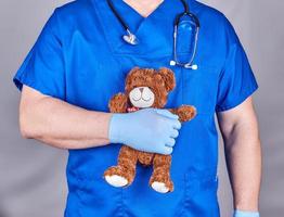 doctor en uniforme azul y viejos guantes de látex sosteniendo un oso de peluche marrón foto