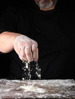 chef en uniforme negro tamiza a través de sus dedos harina de trigo blanco foto
