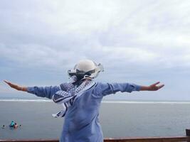 Back of the woman in the hat on the tropical beach who was looking at the sky and the sea while spreading her hands on the bridge. Sea view photo