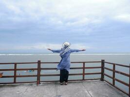 Back of the woman in the hat on the tropical beach who was looking at the sky and the sea while spreading her hands on the bridge. Sea view photo