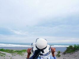 Back of the woman in the hat on the tropical beach who is looking at the sky and the sea while holding her hat photo