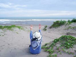 Back of the woman in the hat on the tropical beach who is looking at the sky and the sea while raising her hand in prayer photo