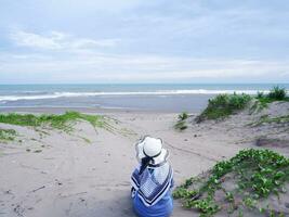 Back of the woman in the hat on the tropical beach who was sitting on the beach sand. background of beach sand and sky. sand mountain.Beach View photo