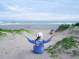 Back of woman in the hat on the tropical beach who was sitting on the sand and looking at the sky and the sea, while spreading her arms . happy women.Beach view photo