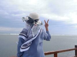 Back of the woman in the hat on the tropical beach who is looking up at the sky and the sea, showing peace gesture. vertical. sea background photo