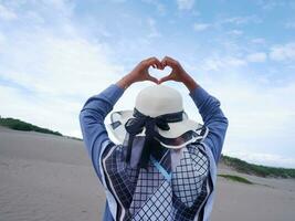 Back of the woman in the hat on the tropical beach who was staring at the ocean then formed a love from the hand. background of beach sand and sky. beach view photo