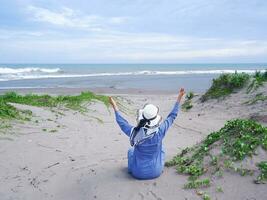 Back of woman in the hat on the tropical beach who was sitting on the sand and looking at the sky and the sea, while spreading her arms . happy women.Beach view photo