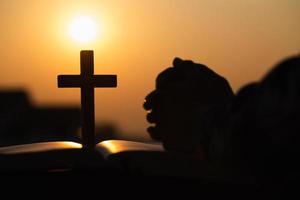 Silhouette of christian young woman praying with a  cross and open the bible at sunrise, Christian Religion concept background. photo