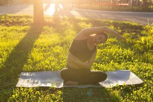 Peaceful young positive pregnant woman in gymnastic suit does yoga and meditate sitting on mat on green grass on sunny warm summer day. Concept of preparation for childbirth and positive attitude photo