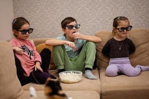 tres niños sentados en la sala de estar, usan gafas 3d viendo películas o dibujos animados. foto