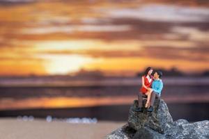 gente en miniatura, pareja sentada en una playa de mar con fondo de puesta de sol foto