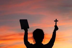 Silhouette of young woman hands holding holy Bible and lift of christian cross with light sunset background. christian concept photo