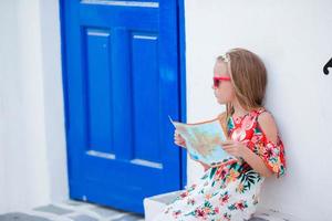 Little girl with map of island outdoors in old streets an Mykonos. Kid at street of typical greek traditional village with white walls and colorful doors on Mykonos Island, in Greece photo