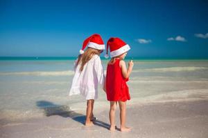 vista trasera de niñas lindas con sombreros de navidad en la playa exótica foto