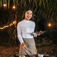 Portrait of Happy Asian young woman cooking meat on barbecue grill at new year party. Bar-B-Q or BBQ on traditional stove. Night Party, people and celebration concept. photo