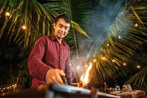 Portrait of Happy Asian young man cooking meat on barbecue grill at new year party. Bar-B-Q or BBQ on traditional stove. Night Party, people and celebration concept. photo