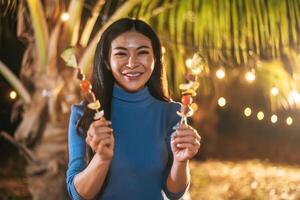 Portrait of Happy Asian young woman cooking meat on barbecue grill at new year party. Bar-B-Q or BBQ on traditional stove. Night Party, people and celebration concept. photo