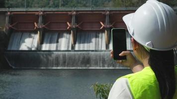 Female engineer in white hat working with smartphone and looking away at dam with hydroelectric power plant and irrigation. video