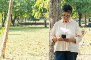 Asian senior woman standing at tree in the park playing on mobile phone photo