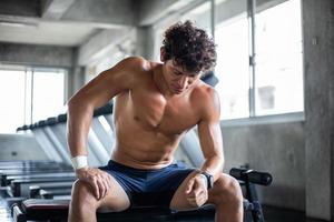 hombre en topless tómese un descanso sentado en el banco del gimnasio después de hacer ejercicio en el gimnasio foto