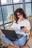 Beautiful businesswoman working on a laptop in a chair near the window. Female freelancer connecting to internet via computer. Blogger or journalist writing new article. photo