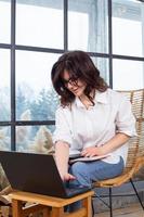 Beautiful businesswoman working on a laptop in a chair near the window. Female freelancer connecting to internet via computer. Blogger or journalist writing new article. photo