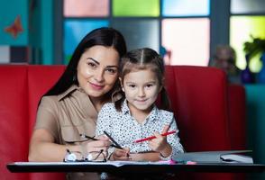 Mother helping her daughter with homework. Portrait of a smiling young mother and her little daughter drawing together in cafe. Happy childhood photo