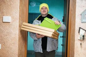 Delivery man with pizza cardboard boxes. Courier in green color hat holding a clipboard while standing against door of residential house photo