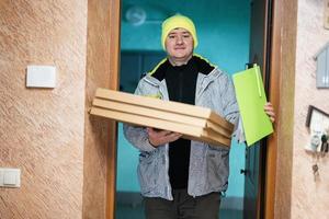 Delivery man with pizza cardboard boxes. Courier in green color hat holding a clipboard while standing against door of residential house photo