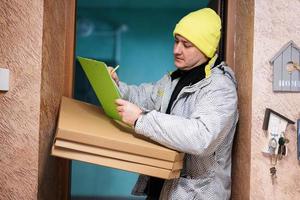 Delivery man with pizza cardboard boxes. Courier in green color hat holding a clipboard while standing against door of residential house photo