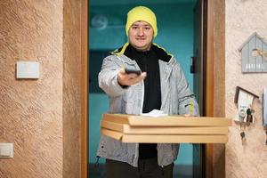 Delivery man with pizza cardboard boxes. Courier in green color hat with mobile phone standing against door of residential house photo